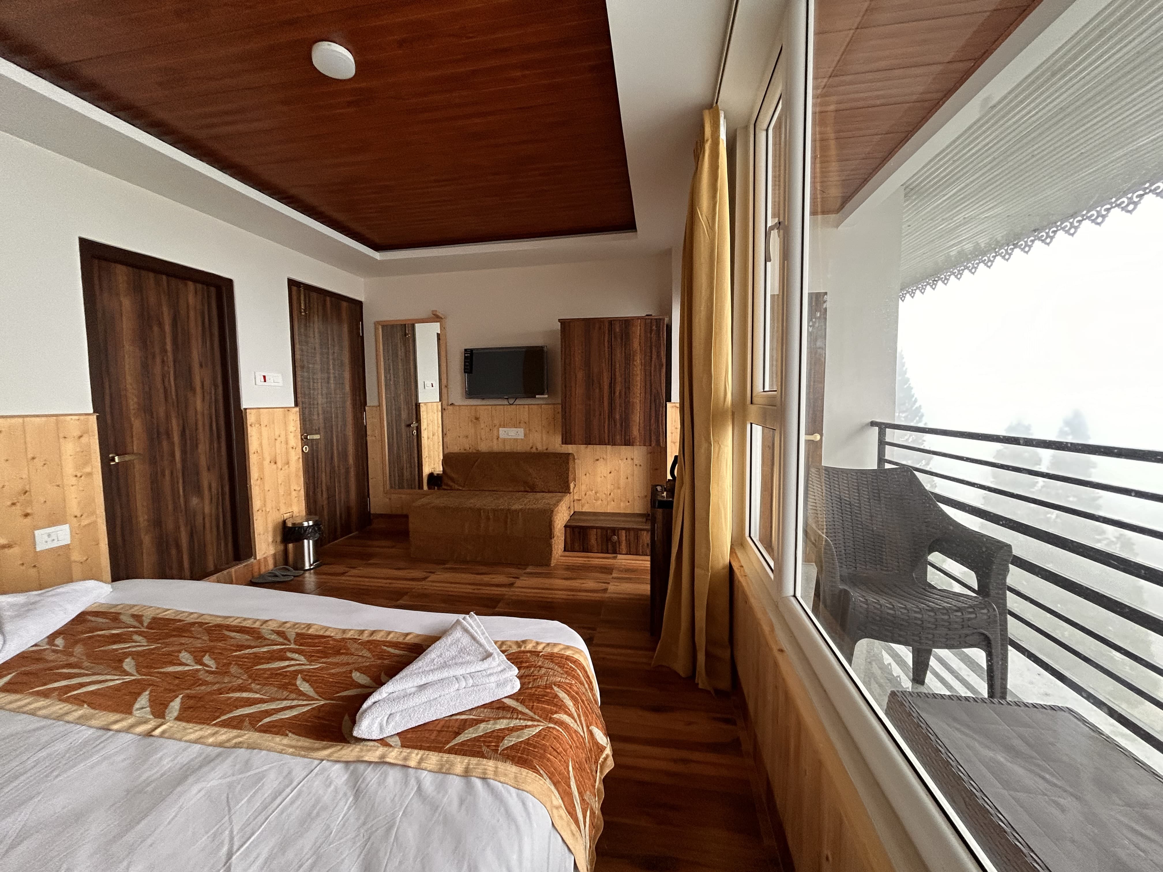 Mountain view room with a wooden ceiling, wall-mounted television and a glazed balcony holding a chair above the tree line